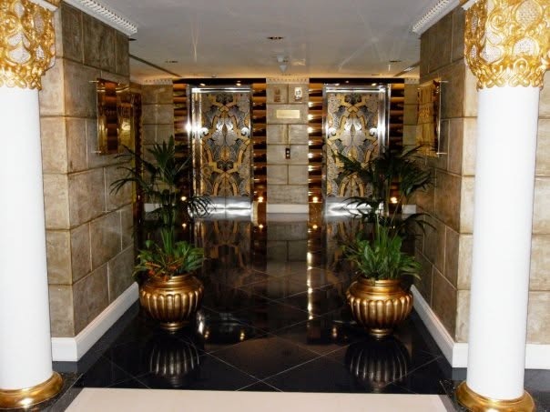 An elevator lobby with gold-patterned doors, black marble floor and gold-capped columns